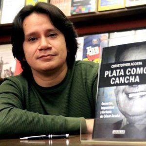 Jornalista peruano Christopher Acosta, de camisa de mangas compridas na cor verde sentado ao lado de seu livro. Ao fundo, vários outros livros. Ele tem cabelos escuros em comprimento médio.