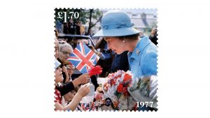 Rainha Elizabeth II veste roupas e chapéu azul, está de lado conversando com o público
