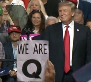 Donald Trump sorrindo em um evento, com um cartaz do QAnon "We Are Q" visível em primeiro plano.