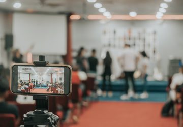 Imagem de uma conferência sendo transmitida online. Em primeiro plano, vemos uma tela de celular filmando o fundo, onde várias pessoas aparecem em uma mesa no centro de um auditório. Eventos híbridos