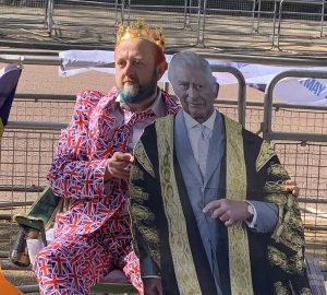 Homem com coroa e terno com bandeira da Inglaterra abraçado a totem do rei Charles III aguardando a passagem do cortejo da coroação no centro de Londres