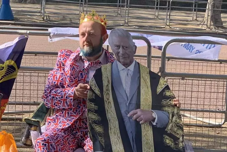 Homem com coroa e terno com bandeira da Inglaterra abraçado a totem do rei Charles III aguardando a passagem do cortejo da coroação no centro de Londres