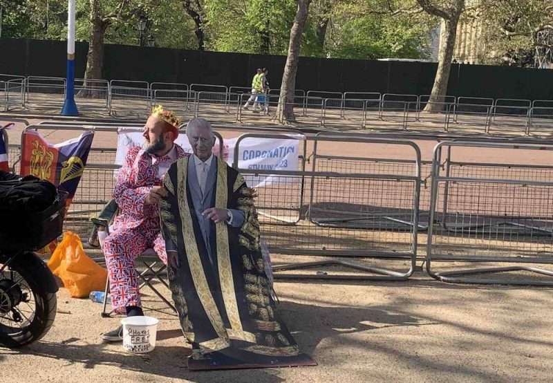 Homem com terno com cores da bandeira inglesa e totem do rei Charles III esperando a passagem do cortejo da coroação