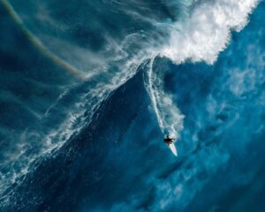 Surfista em uma onda com o arco-íris atrás. A foto foi premiada no concurso de fotografia dos oceanos