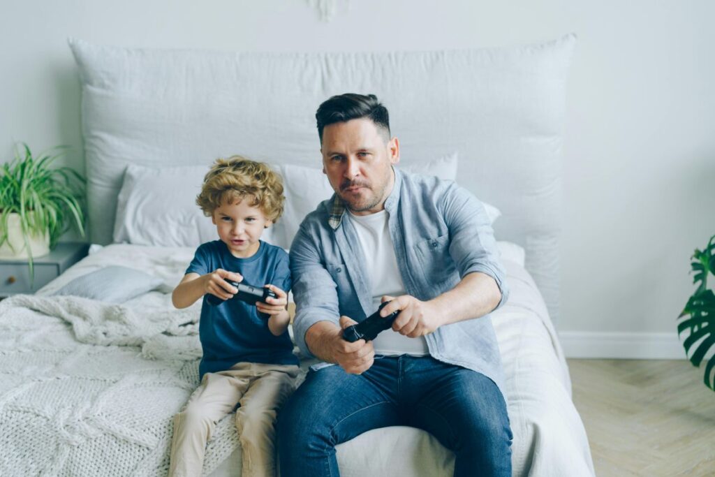 Pai e filho jogam videogame sentados na cama; estudo aponta que jogos podem ajudar a diminuir o estresse em curto prazo