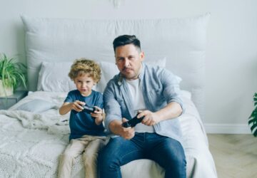 Pai e filho jogam videogame sentados na cama; estudo aponta que jogos podem ajudar a diminuir o estresse em curto prazo