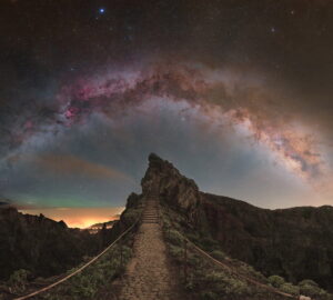 Via Láctea fotografada em arco sobre colina rochosa na Ilha da Madeira, Portugal, com nebulosas vermelhas visíveis.