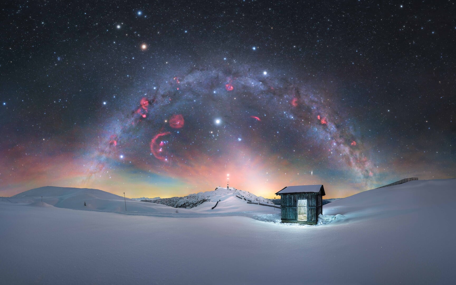 Via Láctea de inverno com nebulosas vermelhas sobre o Parque Natural Dobratsch, Áustria, com cabana nevada em primeiro plano.