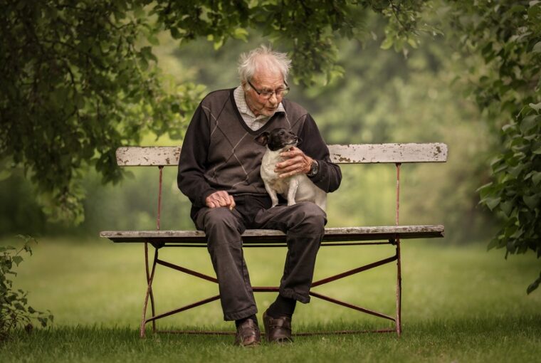 Homem sentado em um banco em um jardim com um cão no colo. A foto foi premiada no Dog Photography Awards