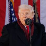 Donald Trump fazendo discurso com bandeiras ao fundo