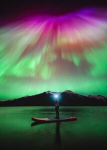 As luzes do norte iluminando o lago e uma pessoa em um stand up paddle, no Canadá