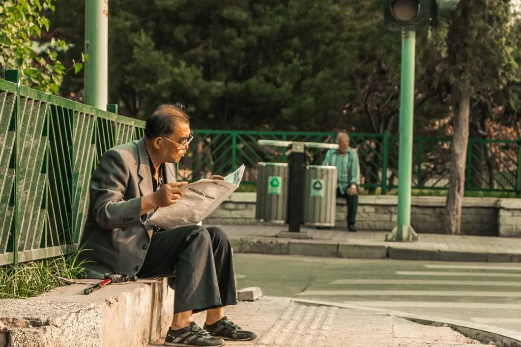 Homem lendo jornal em parque com latas de reciclagem ao fundo