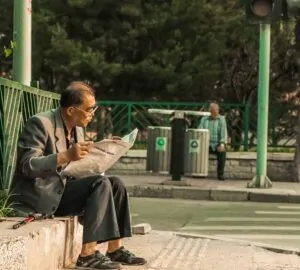 Homem lendo jornal em parque com latas de reciclagem ao fundo