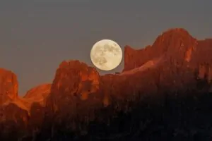 A Lua cheia surge sobre as montanhas Dolomitas, tingidas pelo dourado do pôr do sol. 