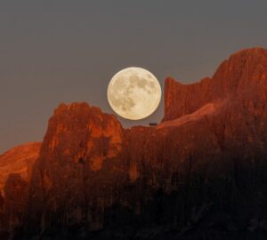 A Lua cheia surge sobre as montanhas Dolomitas, tingidas pelo dourado do pôr do sol.