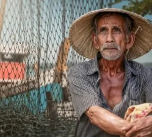 Pescador idoso com rede e barco ao fundo em cenário de pesca tradicional