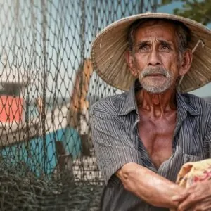 Pescador idoso com rede e barco ao fundo em cenário de pesca tradicional