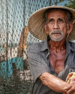 Pescador idoso com rede e barco ao fundo em cenário de pesca tradicional