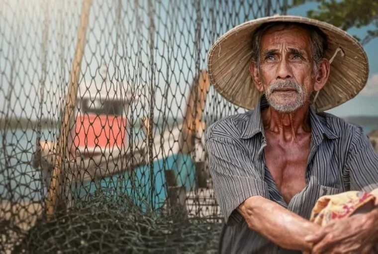 Pescador idoso com rede e barco ao fundo em cenário de pesca tradicional