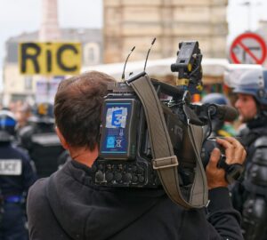 Cinegrafista em protesto com policiais ao fundo - cobertura livre de manifestações é uma das ameaças a jornalistas listadas pela ONU
