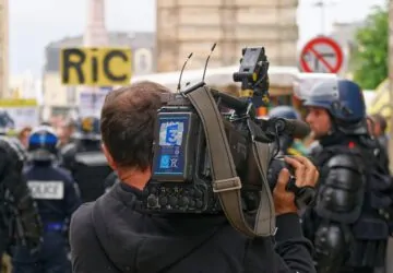 Cinegrafista em protesto com policiais ao fundo - cobertura livre de manifestações é uma das ameaças a jornalistas listadas pela ONU