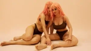 Duas mulheres ruivas de biquine preto sentadas em um fundo rosa. 