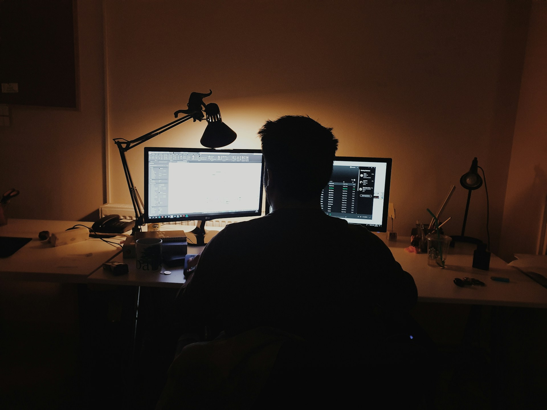Homem de costas em sala escura trabalhando em dois computadores