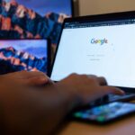 Pessoa digitando em um notebook na página inicial do Google, com um celular ao lado das mãos e um tablet no fundo