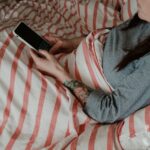Mulher jovem deitada na cama sob cobertor usando smartphone