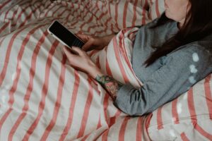 Mulher jovem deitada na cama sob cobertor usando smartphone