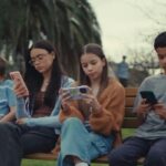Crianças sentadas em um banco de praça com o celular nas mãos.