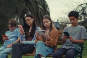 Crianças sentadas em um banco de praça com o celular nas mãos.